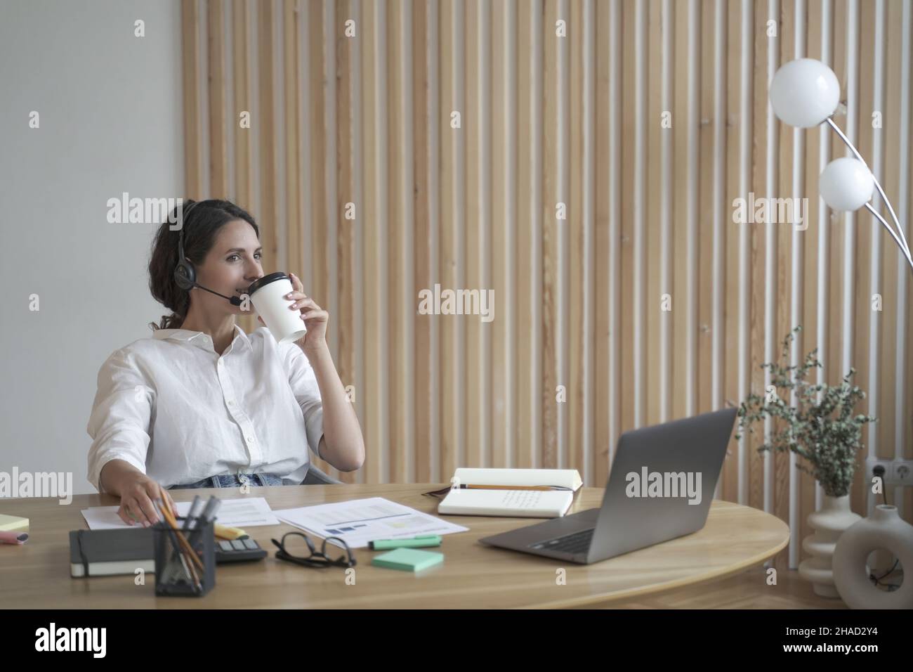 Soddisfatto positivo spagnolo imprenditore femminile in cuffia wireless bere caffè in ufficio Foto Stock