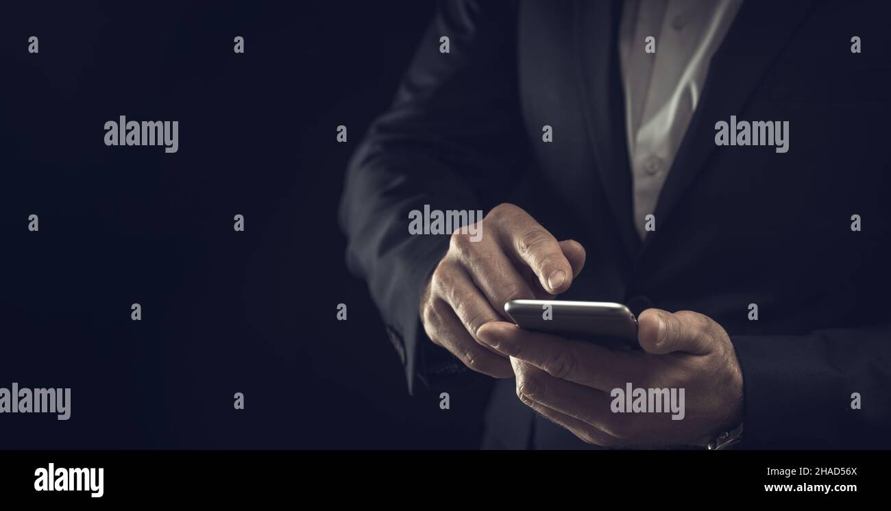 Un uomo d'affari in abito scuro e una camicia bianca, su sfondo nero, con uno smartphone nelle sue mani e un posto per il tuo testo. Foto Stock