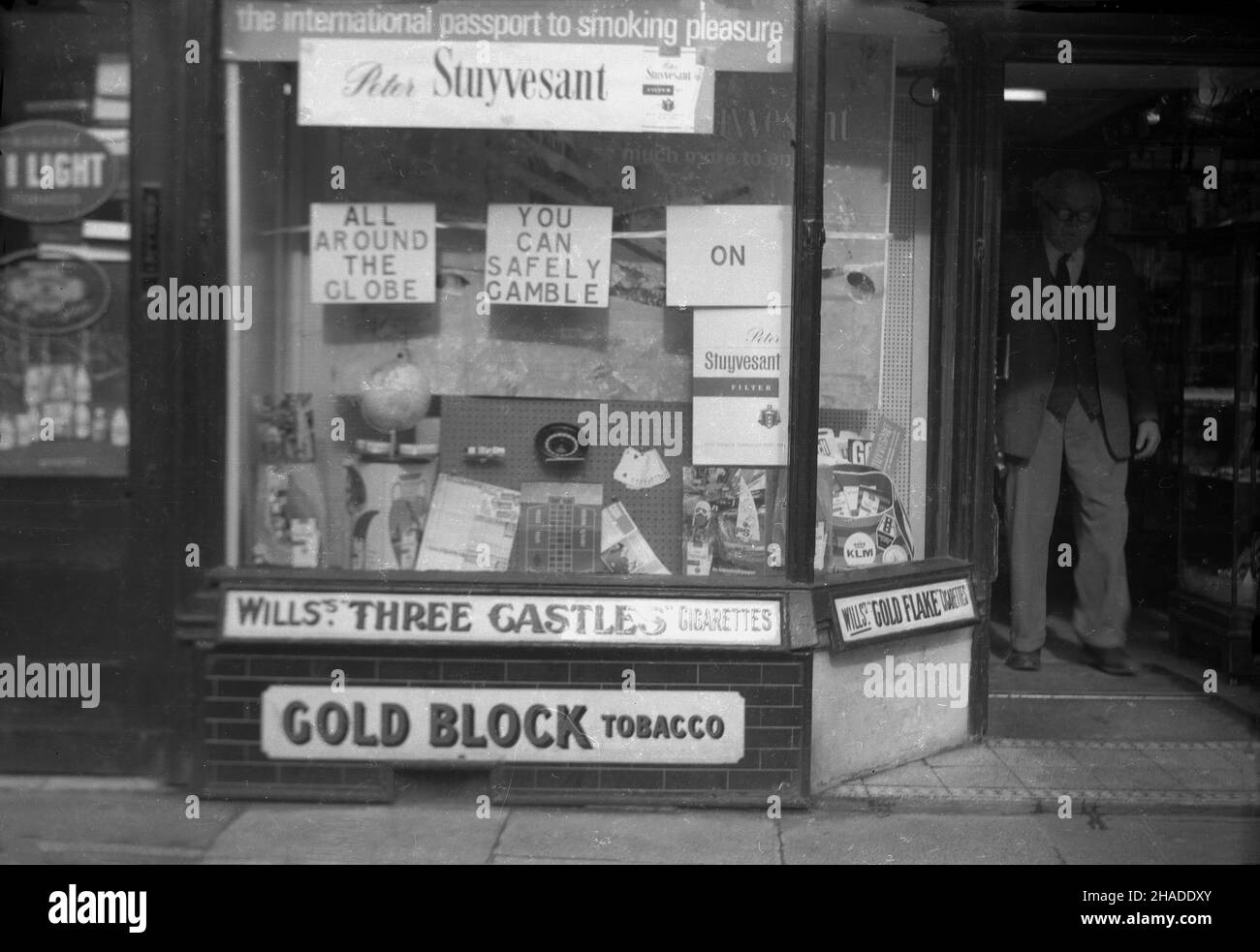 1950s, storico, negoziante maschile alla porta del suo tabacconista, John Isaacs in Half Moon Street, Sherborne, Dorset, Inghilterra, Regno Unito. Nella finestra, la pubblicità per le nuove sigarette Peter Stuyvesant, 'il passaporto internazionale per il piacere del fumo e 'tutto il Globe, si può tranquillamente scommettere su Peter Stuyvesant, Foto Stock