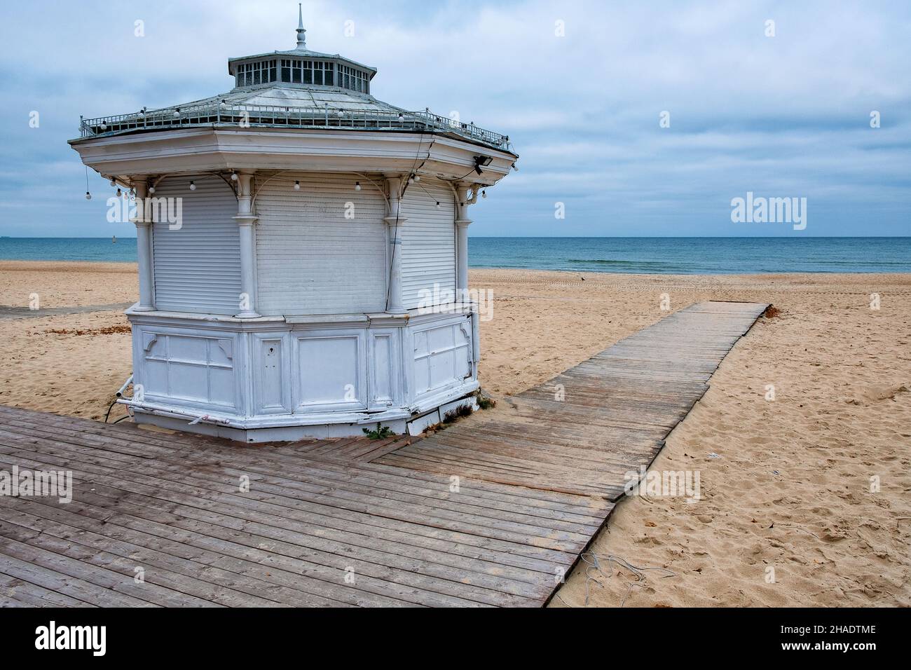 Una spiaggia vacanziera vuota e chiusa in autunno, rampa di legno che porta verso l'acqua con una capanna bianca chiusa dopo la stagione e cielo nuvoloso Foto Stock