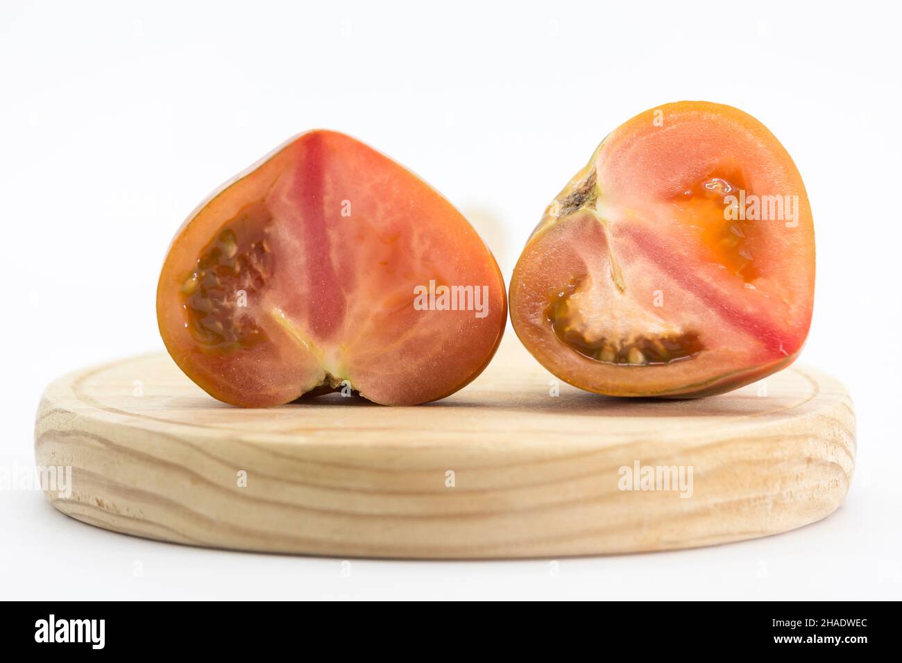 pomodoro tagliato a metà e posto su un asse di legno rotondo, tutto su sfondo bianco Foto Stock