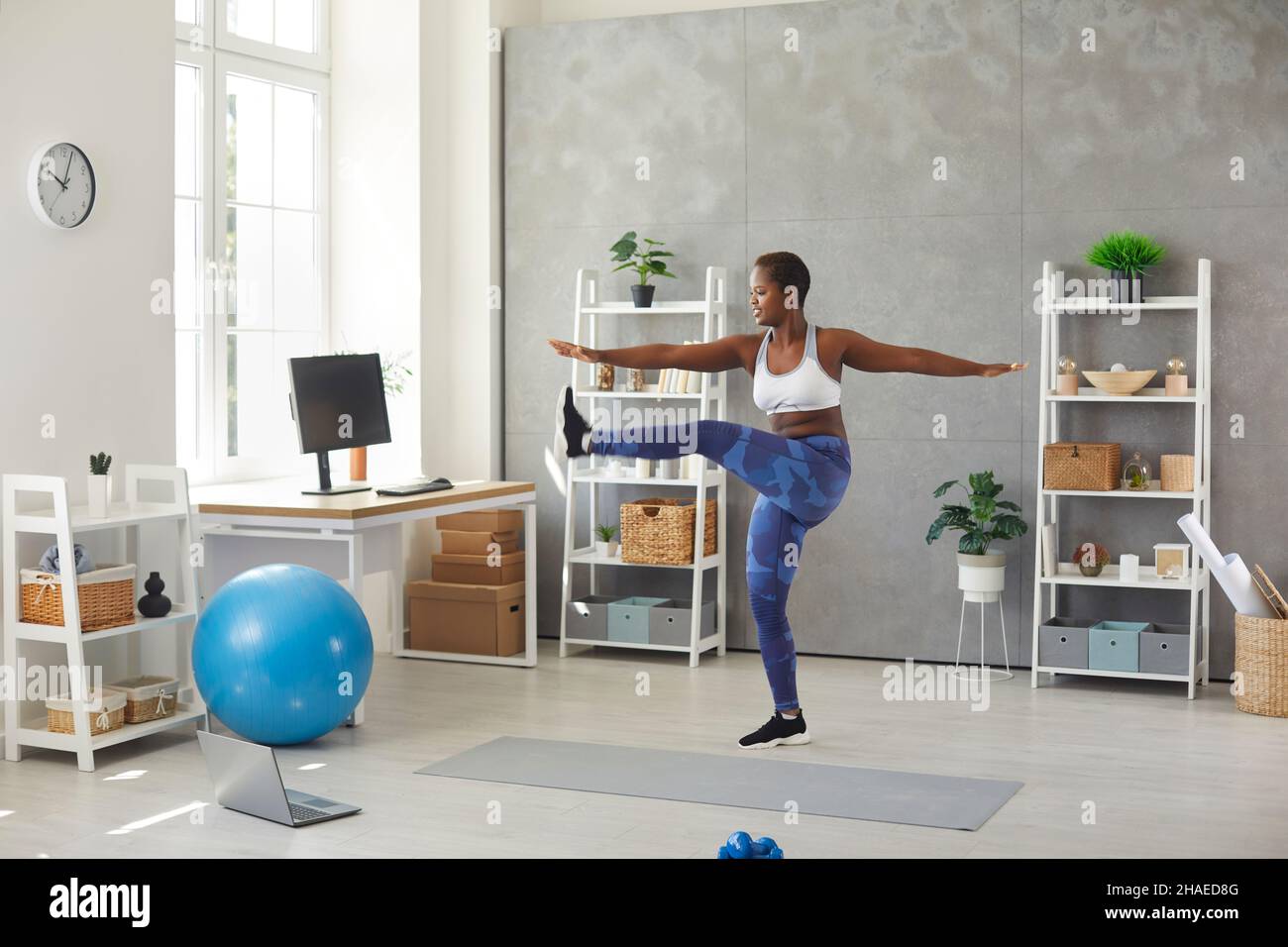 Donna nera che fa le oscillazioni della gamba a casa guardando la lezione di allenamento online di fitness sul laptop Foto Stock