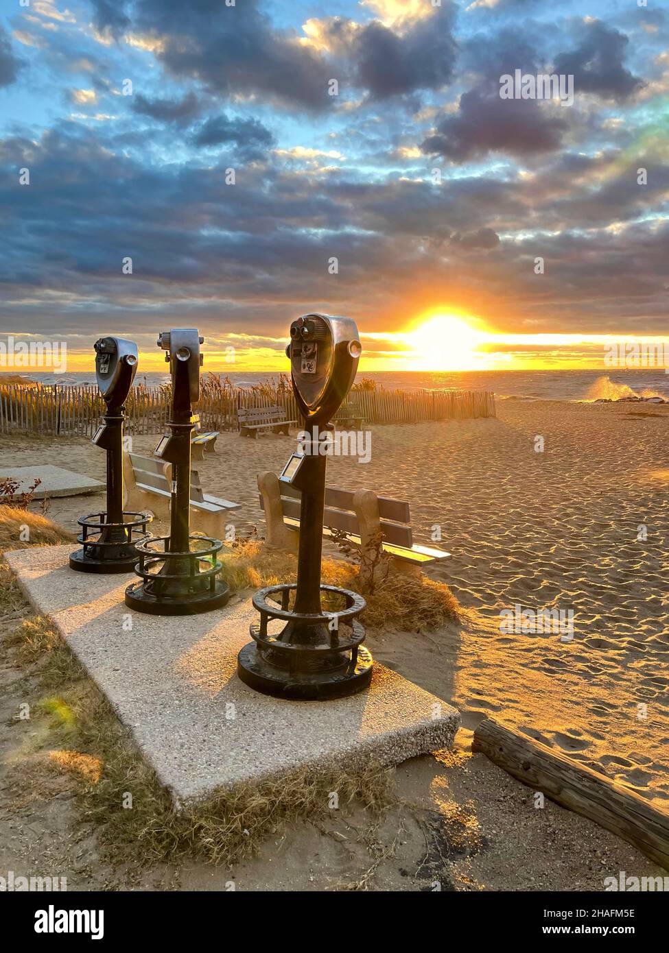 Tre serie di binocoli a gettoni in piedi su una spiaggia al tramonto con onde che si infrangono e il sole basso sullo sfondo. Foto Stock