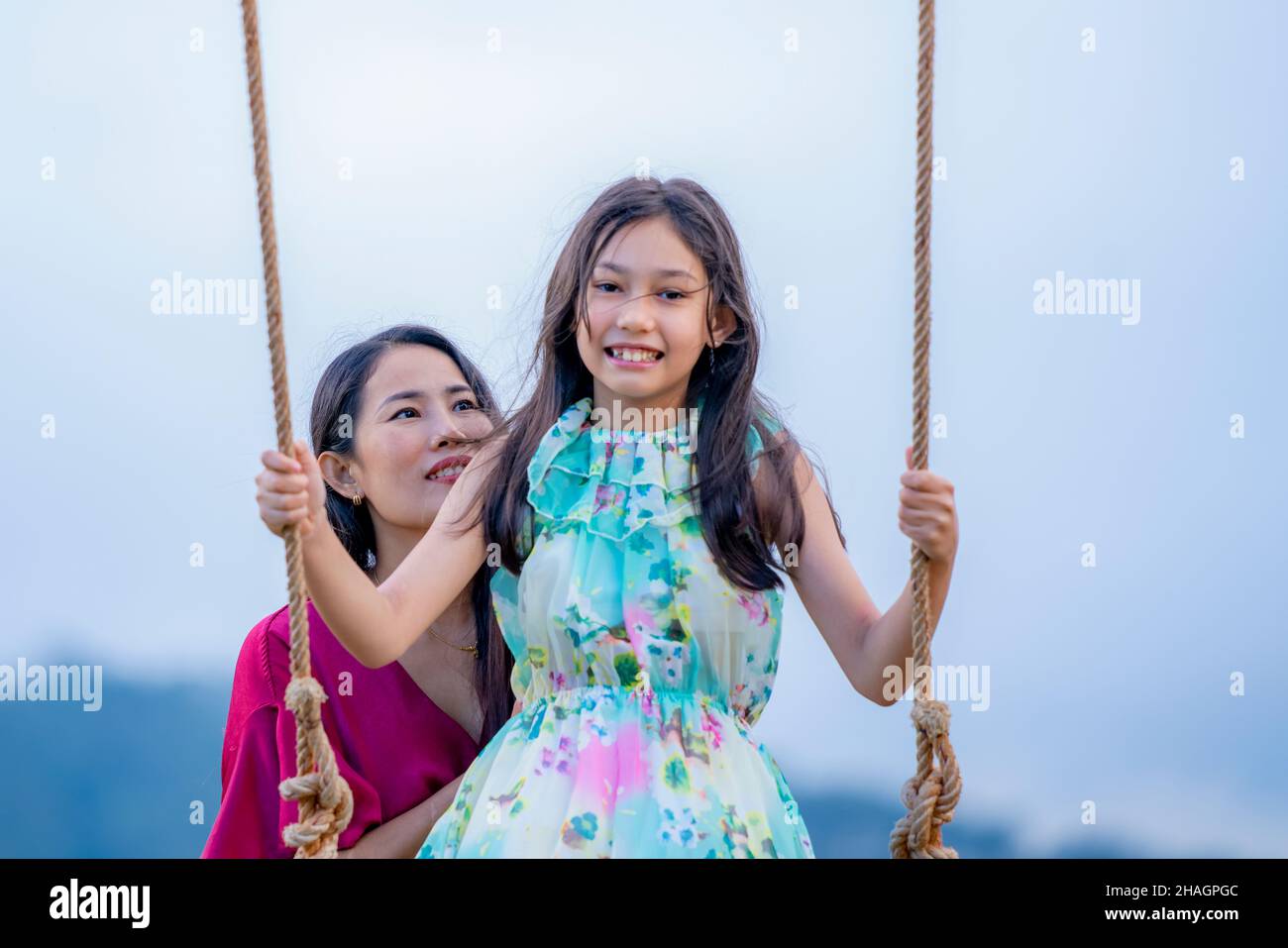 Fuoco a Mom.Happy ragazza che oscilla giocoso su un swing. Madre e figlia si godono la giornata estiva in un parco giochi sopra i fiori gialli montagna. Foto Stock