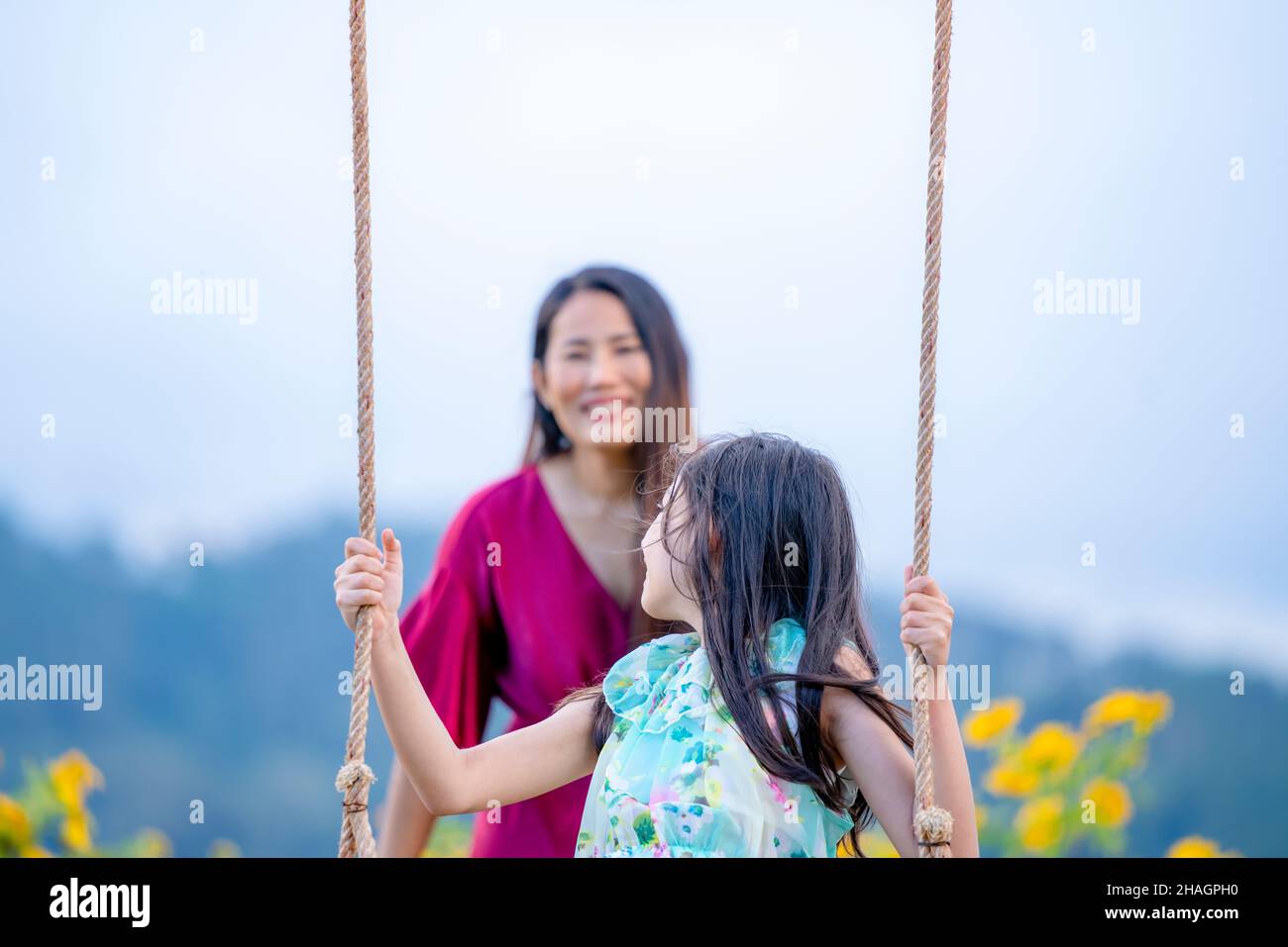 Ragazza felice che oscillano giocoso su un'oscillazione. Madre e figlia si godono la giornata estiva in un parco giochi sopra i fiori gialli montagna. Foto Stock