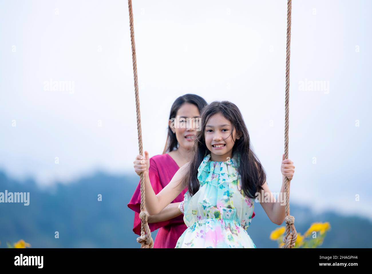 Ragazza felice che oscillano giocoso su un'oscillazione. Madre e figlia si godono la giornata estiva in un parco giochi sopra i fiori gialli montagna. Foto Stock