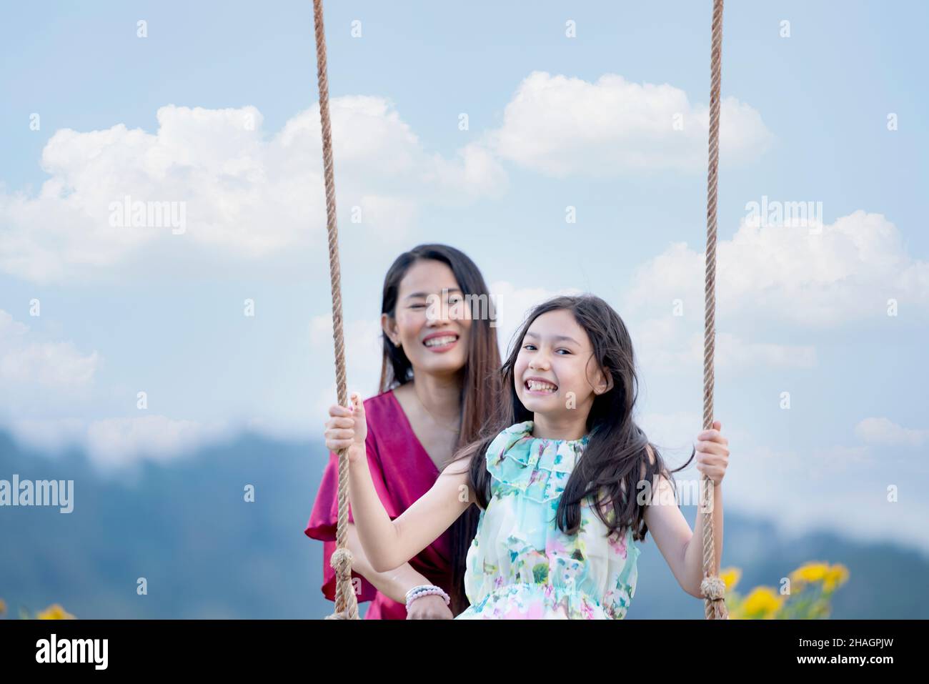Ragazza felice che oscillano giocoso su un'oscillazione. Madre e figlia si godono la giornata estiva in un parco giochi sopra i fiori gialli montagna. Foto Stock