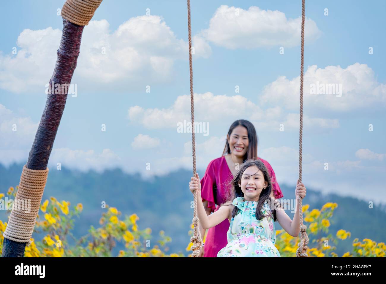 Ragazza felice che oscillano giocoso su un'oscillazione. Madre e figlia si godono la giornata estiva in un parco giochi sopra i fiori gialli montagna. Foto Stock