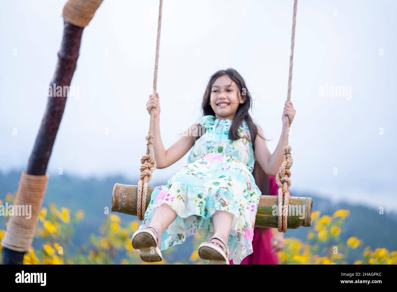 Sfocatura astratta di ragazza felice che oscilla giocoso su un swing. Madre e figlia si godono la giornata estiva in un parco giochi sopra i fiori gialli montagna. Foto Stock