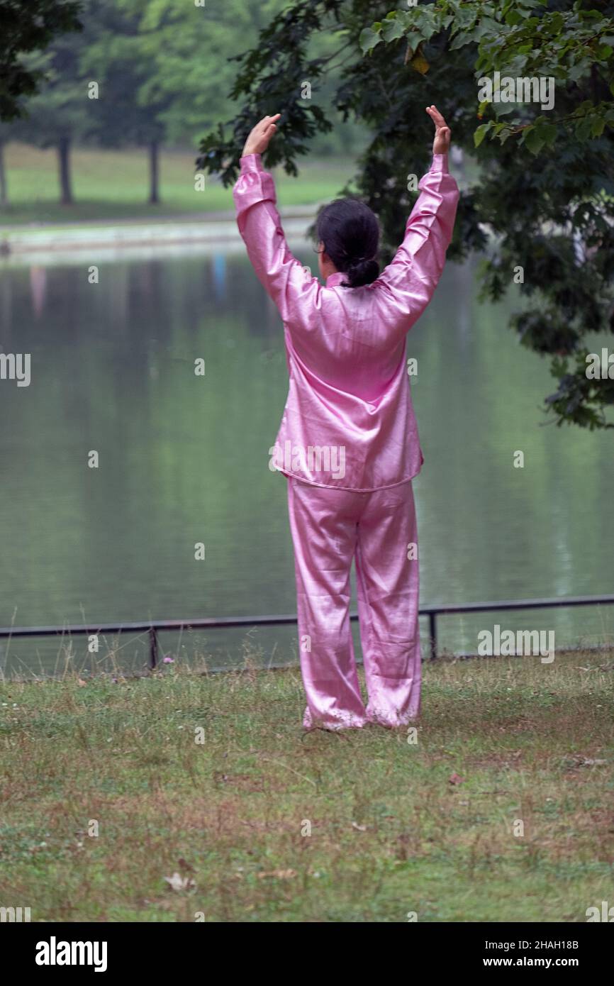 Una donna in una tuta Tai Chi rosa fa esercizi presso il lago in un parco a Flushing, Queens, New York. Foto Stock