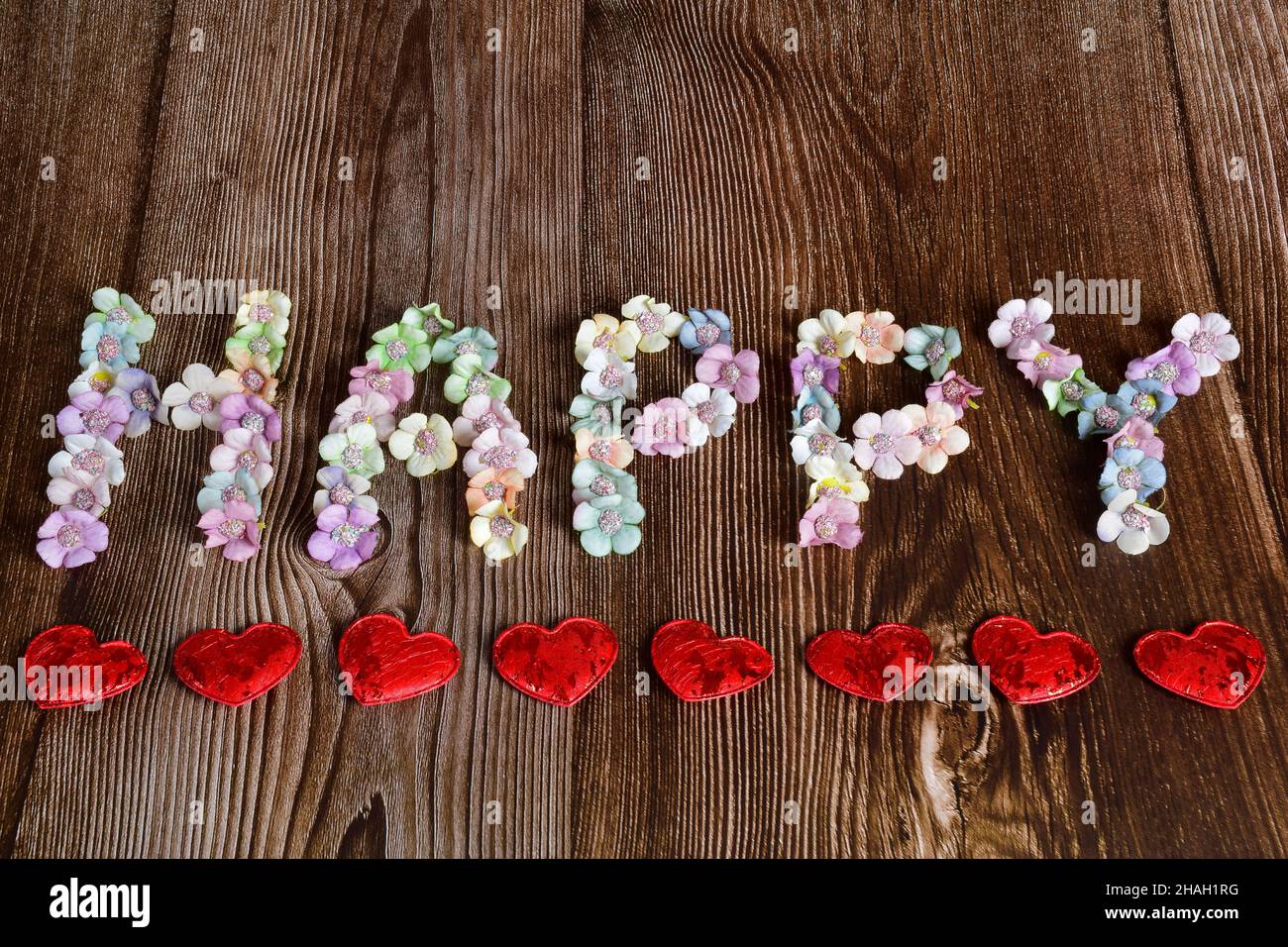 Su uno sfondo di legno marrone, la parola FELICE è scritto in grandi lettere di fiori multicolore. I cuori decorativi sono allineati da sotto in a l Foto Stock