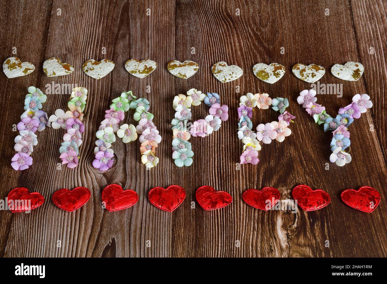Su uno sfondo di legno marrone, la parola FELICE è scritto in grandi lettere di fiori multicolore. I cuori decorativi sono allineati superiore e inferiore in un Foto Stock