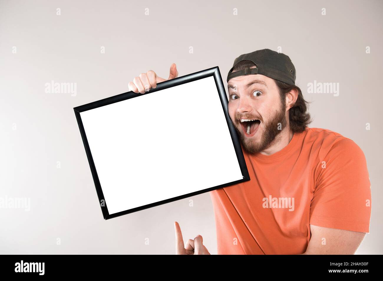 Giovane uomo bearded che fa faccia divertente, tenendo un bordo vuoto del segno. Studio girato su sfondo grigio. Foto Stock