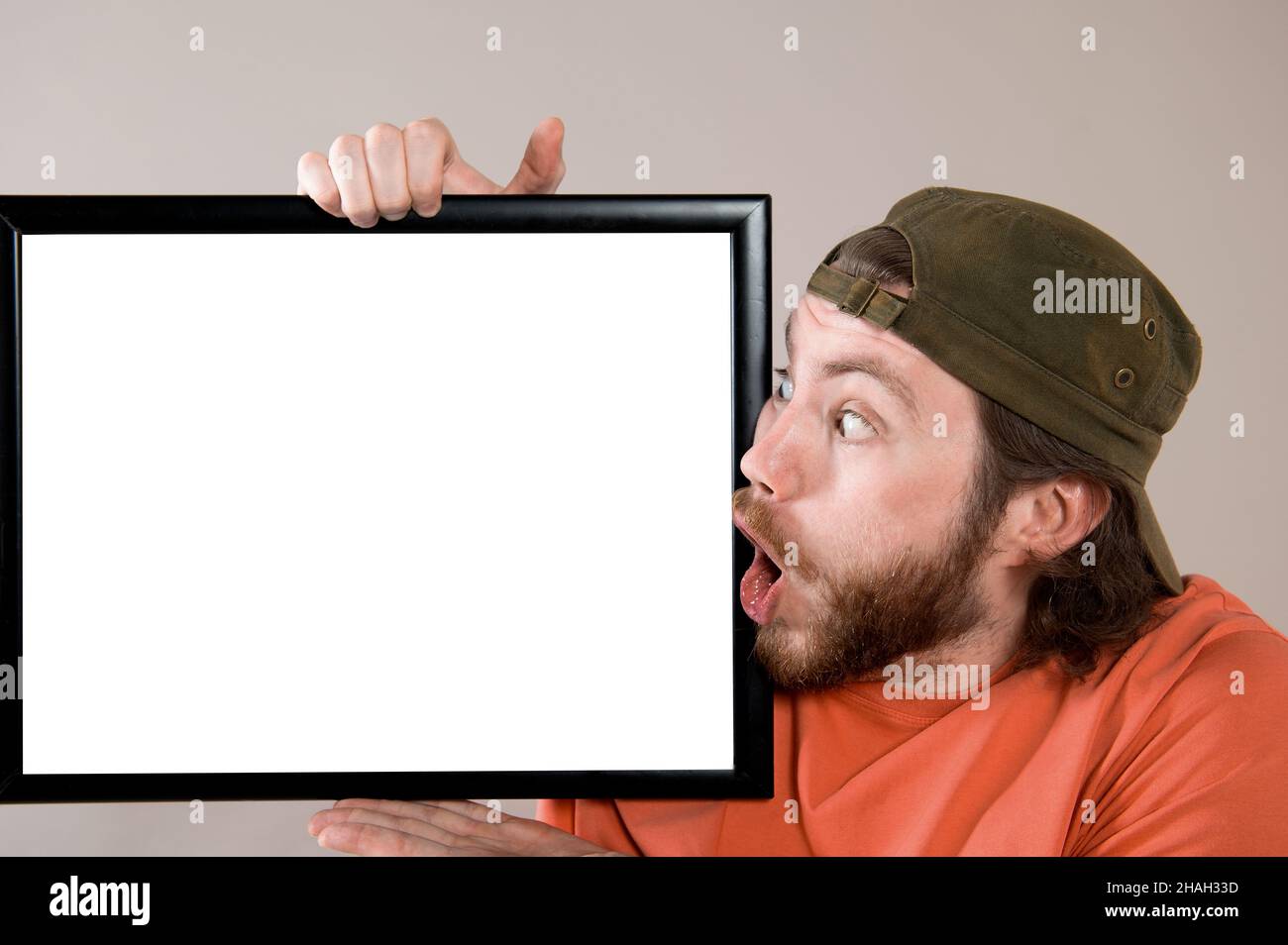 Giovane uomo bearded che fa faccia divertente, tenendo un bordo vuoto del segno. Studio girato su sfondo grigio. Foto Stock