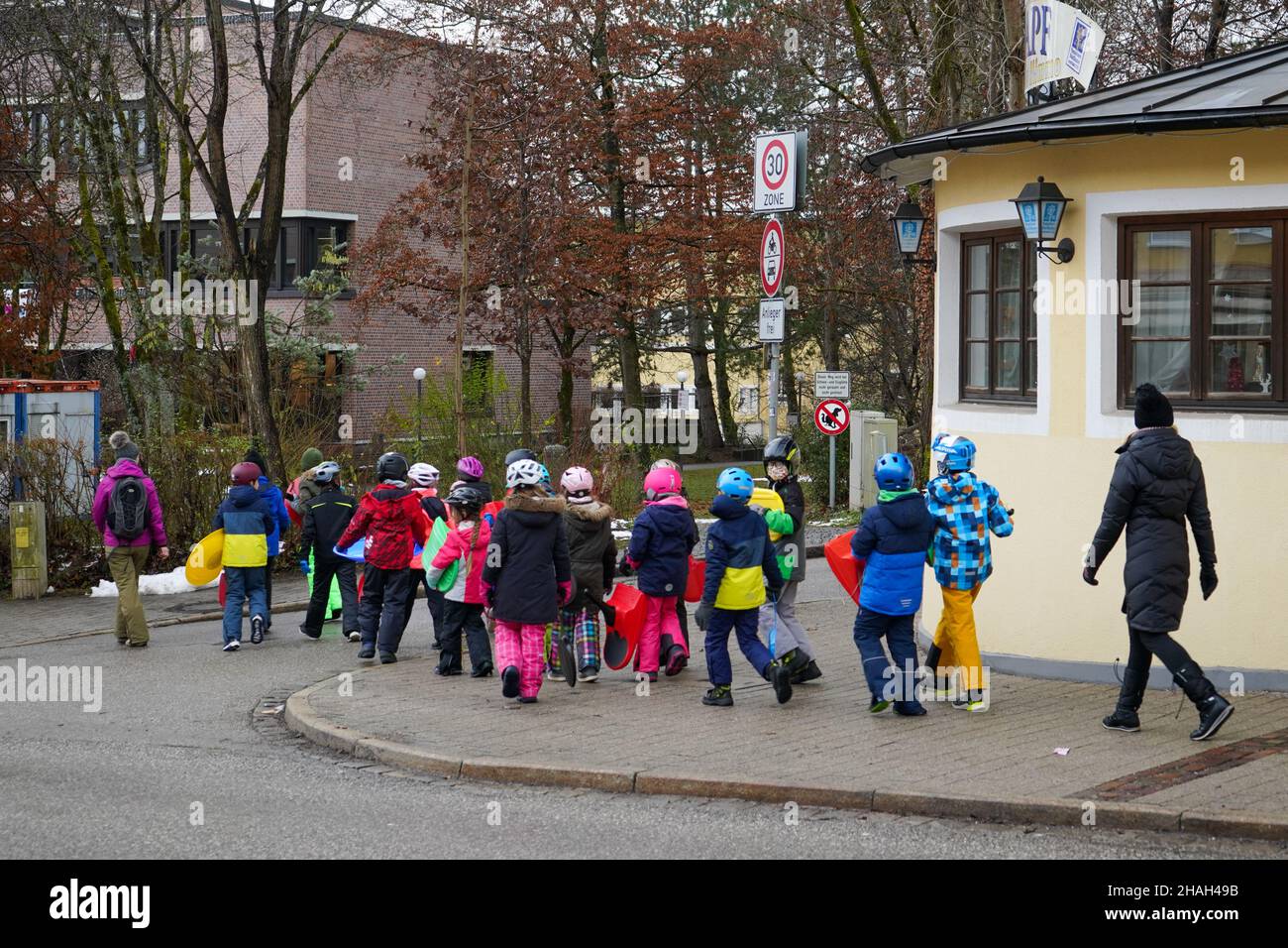 Un gruppo di bambini che indossa slitte da neve in plastica e attraversa una strada con i loro vestiti colorati davanti e dietro un educatore per la sicurezza. Foto Stock