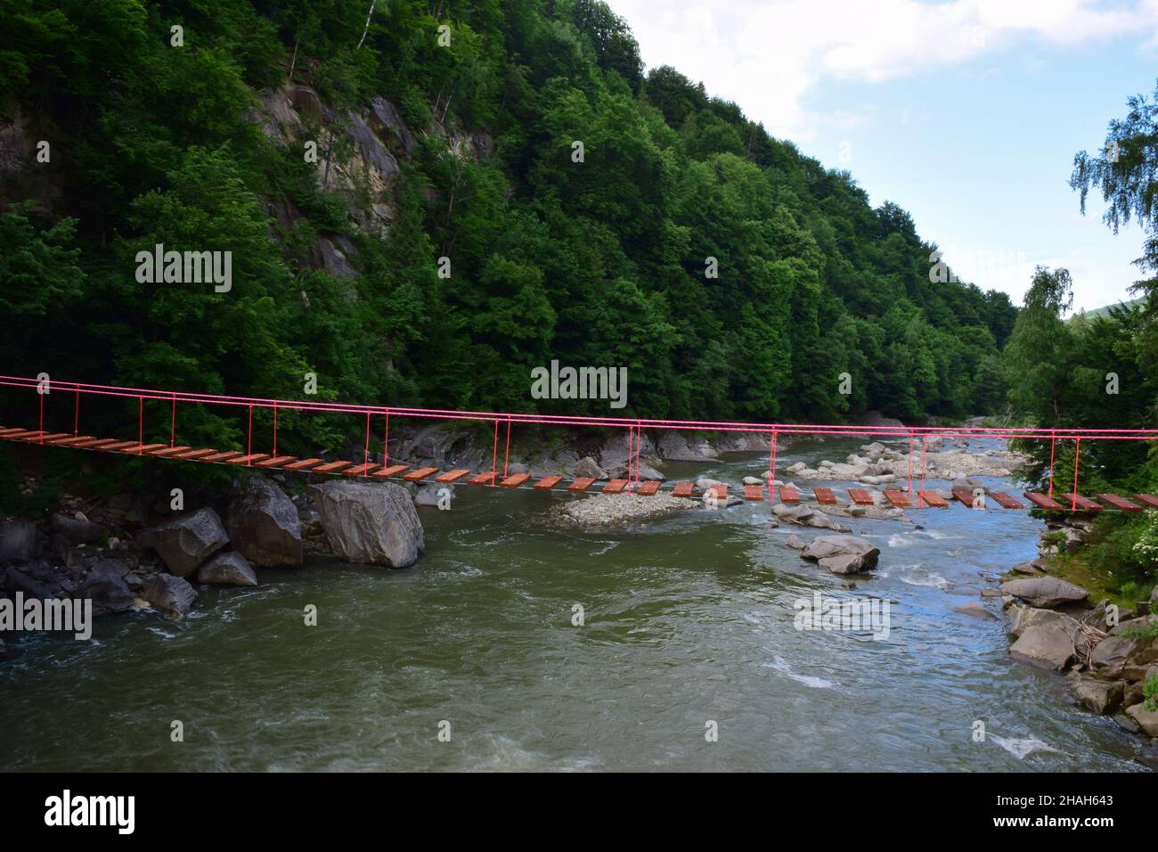 Ponte sospeso a corda senza persone tra le rive rocciose su un fiume di montagna sullo sfondo di una montagna in vegetazione lussureggiante Foto Stock