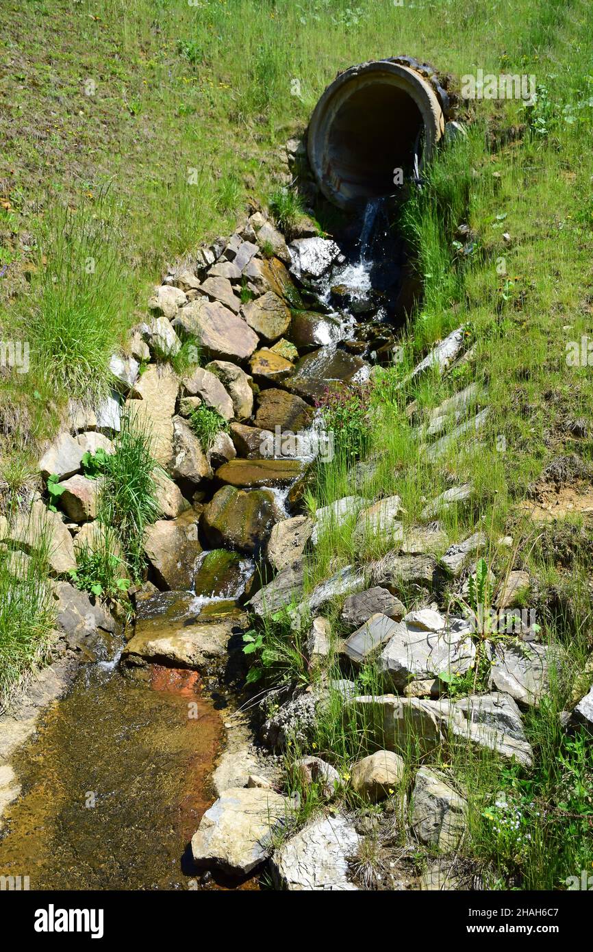 Da un grande tubo di calcestruzzo nella collina, una sorgente con acqua pulita defluisce e scorre lungo una grondaia appositamente disposta in pietra. Foto Stock