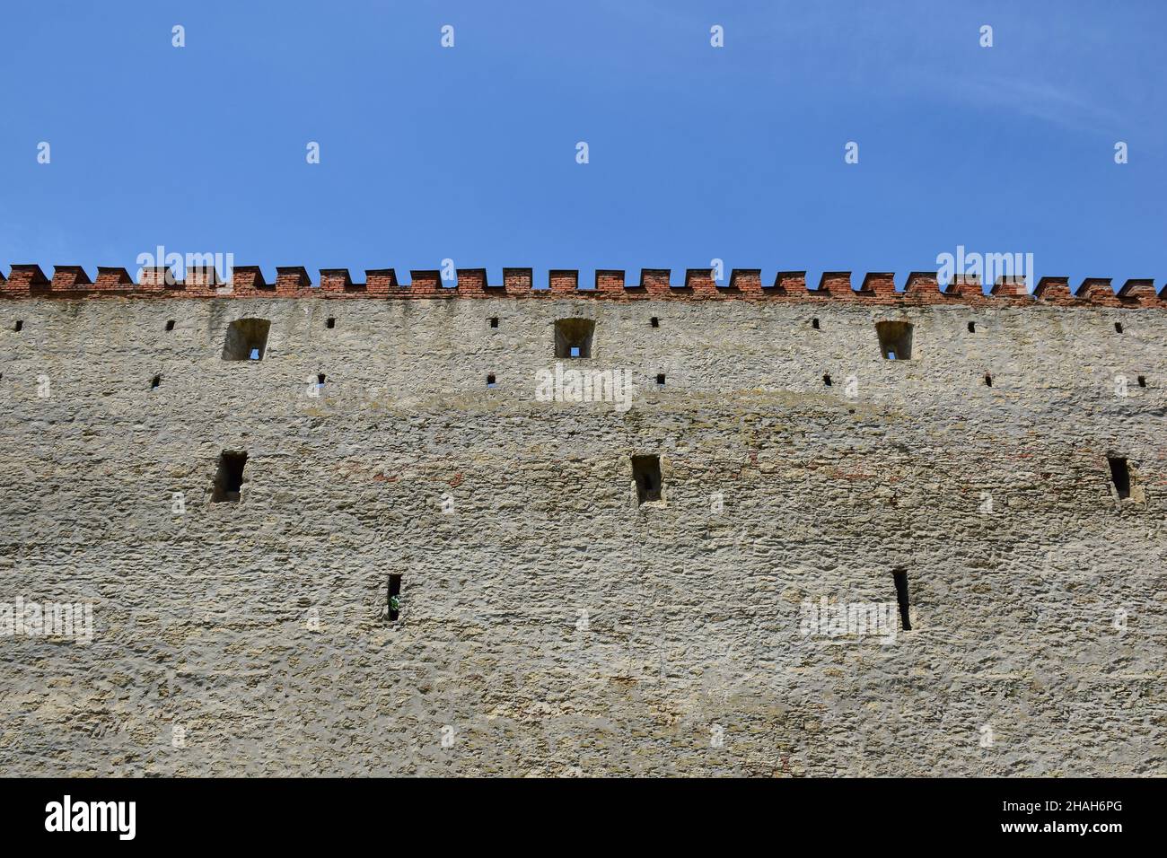 Muro di pietra medievale di un castello o fortezza con cime di mattoni frastagliate. Ci sono piccole scappatoie per sparare. Sopra il cielo blu Foto Stock