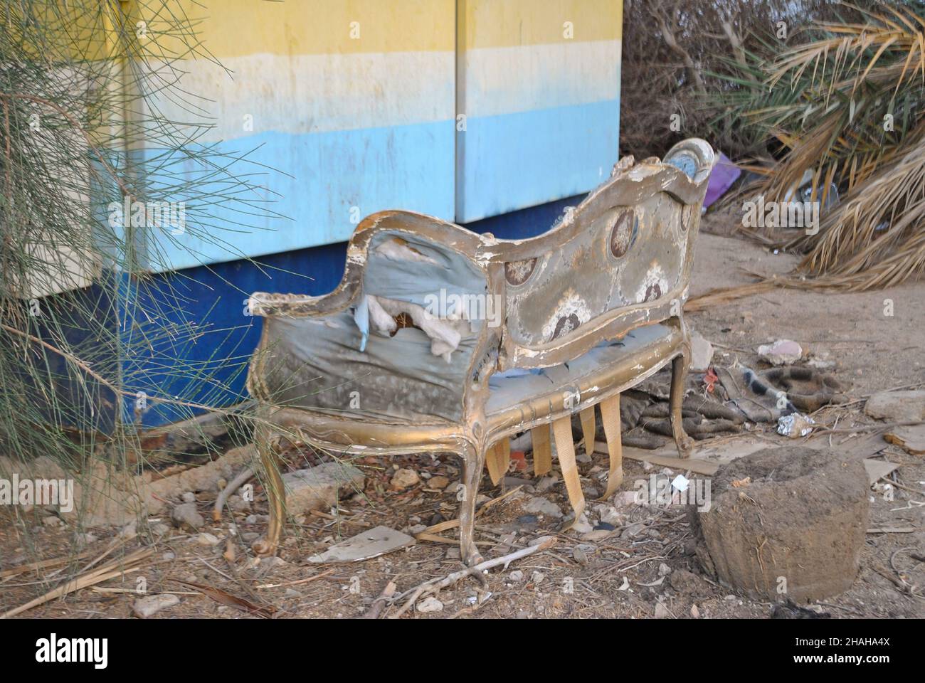 Un vecchio divano sciablato e lacerato con i suoi lati che cadono fuori siede in un deposito vicino ad un mucchio di rifiuti e ad un muro dipinto Foto Stock