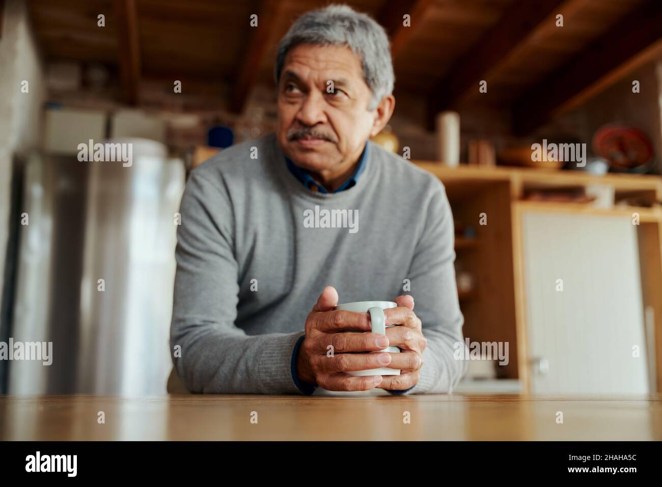 Pensive pensionato biraciale anziano maschio appoggiandosi sul banco della cucina moderna, tenendo il caffè del mattino nelle sue mani. Foto Stock