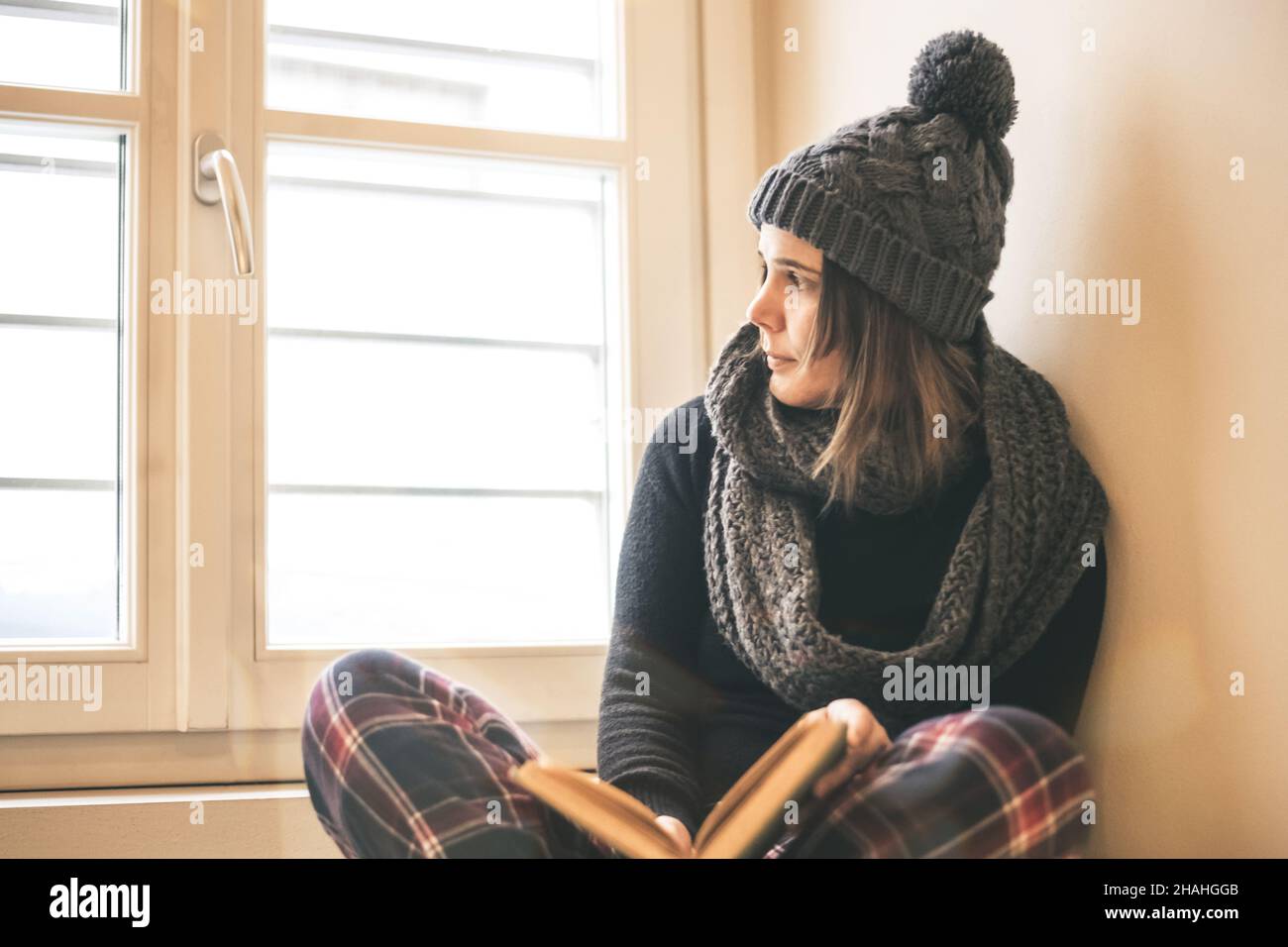Giovane bella ragazza che legge un libro durante l'inverno freddo con  cappellino di lana e sciarpa per mantenere caldo. Donna seduta leggere  vicino alla finestra. Donna che si rilassa a casa Foto