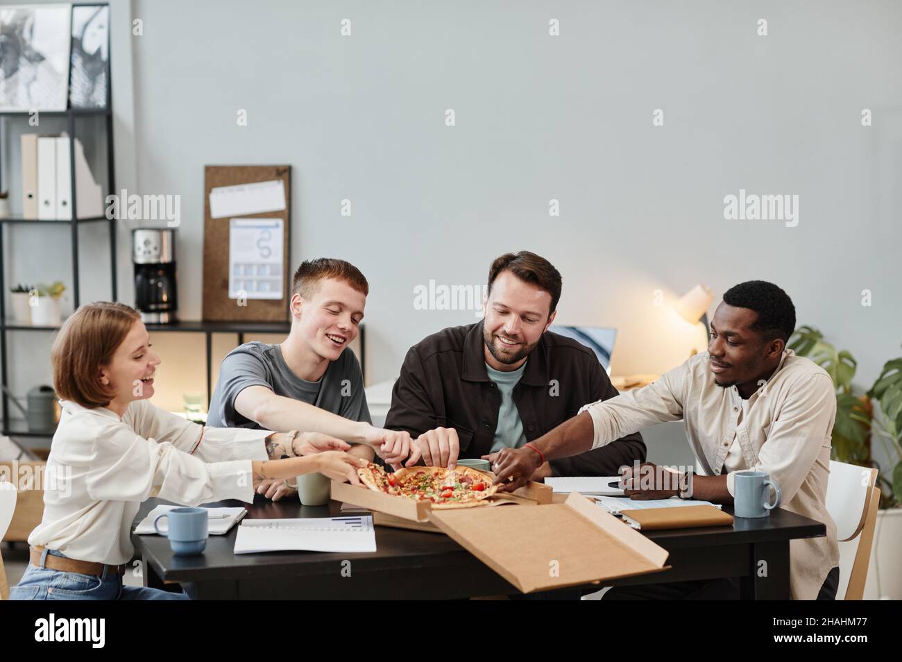 I giovani uomini d'affari ordinano la pizza per il pranzo in ufficio, si siedono al tavolo e si gustano il loro pasto Foto Stock