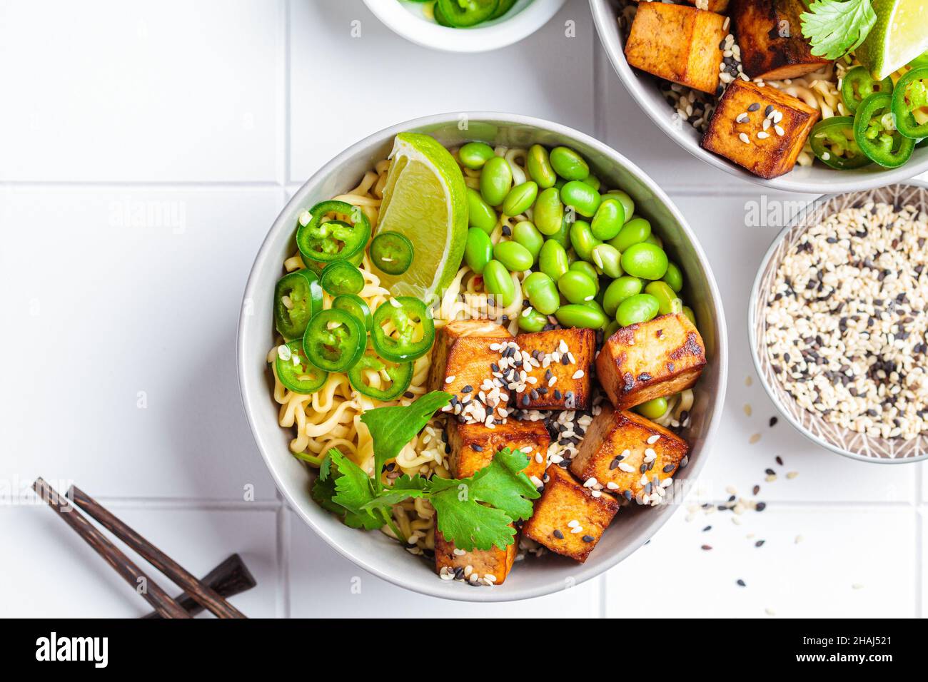 Zuppa di ramen di tagliatelle vegane con tofu marinato, fagioli edamame e peperoni caldi in una ciotola grigia su sfondo bianco piastrellato, vista dall'alto. Foto Stock