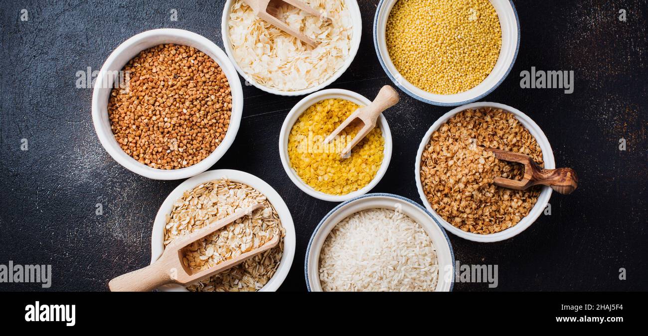 Varietà di cereali fiocchi di riso, miglio, grano saraceno, farinata d'avena. Superfood in ciotole di ceramica bianca su sfondo di cemento vecchio scuro. Vista dall'alto. Foto Stock