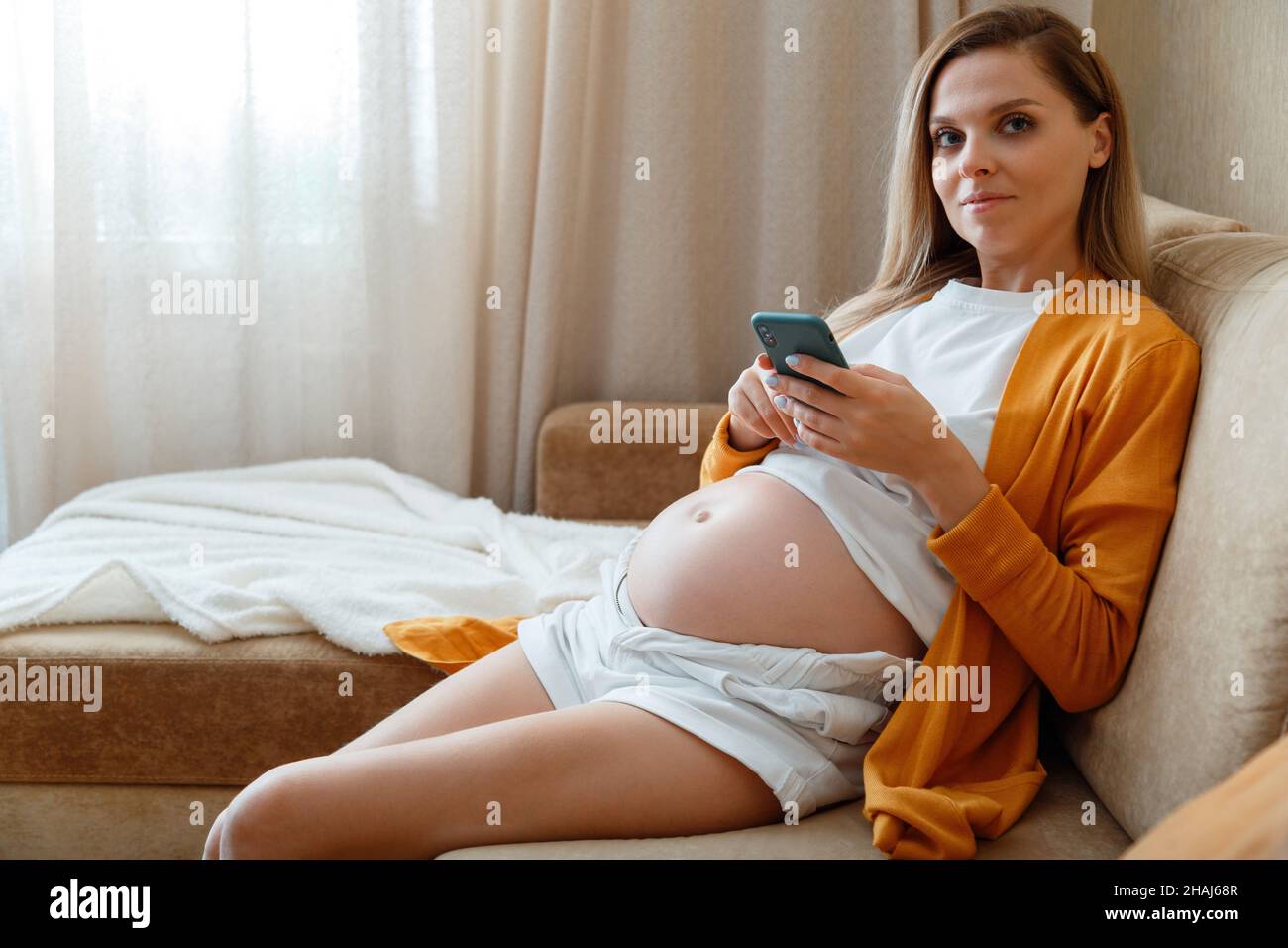 Ritratto di giovane donna incinta di 30 s seduta sul divano in casa accogliente. Donna incinta caucasica bionda con pancia utilizzare smartphone per Foto Stock