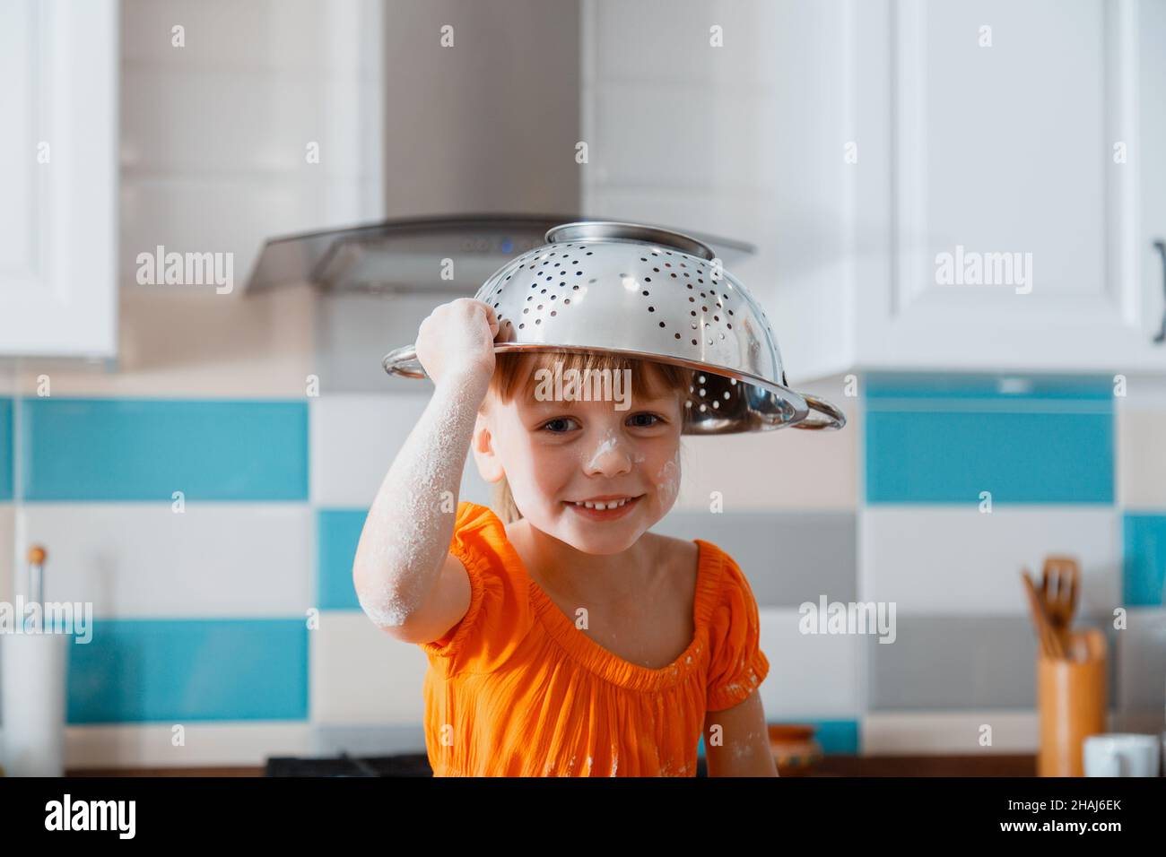 Felice ritratto della figlia che gioca in cucina. Bambino ragazza capretto sporcato in farina mettere colander ciotola sulla testa come cappello hanno divertimento e risate gioco. Felice Foto Stock