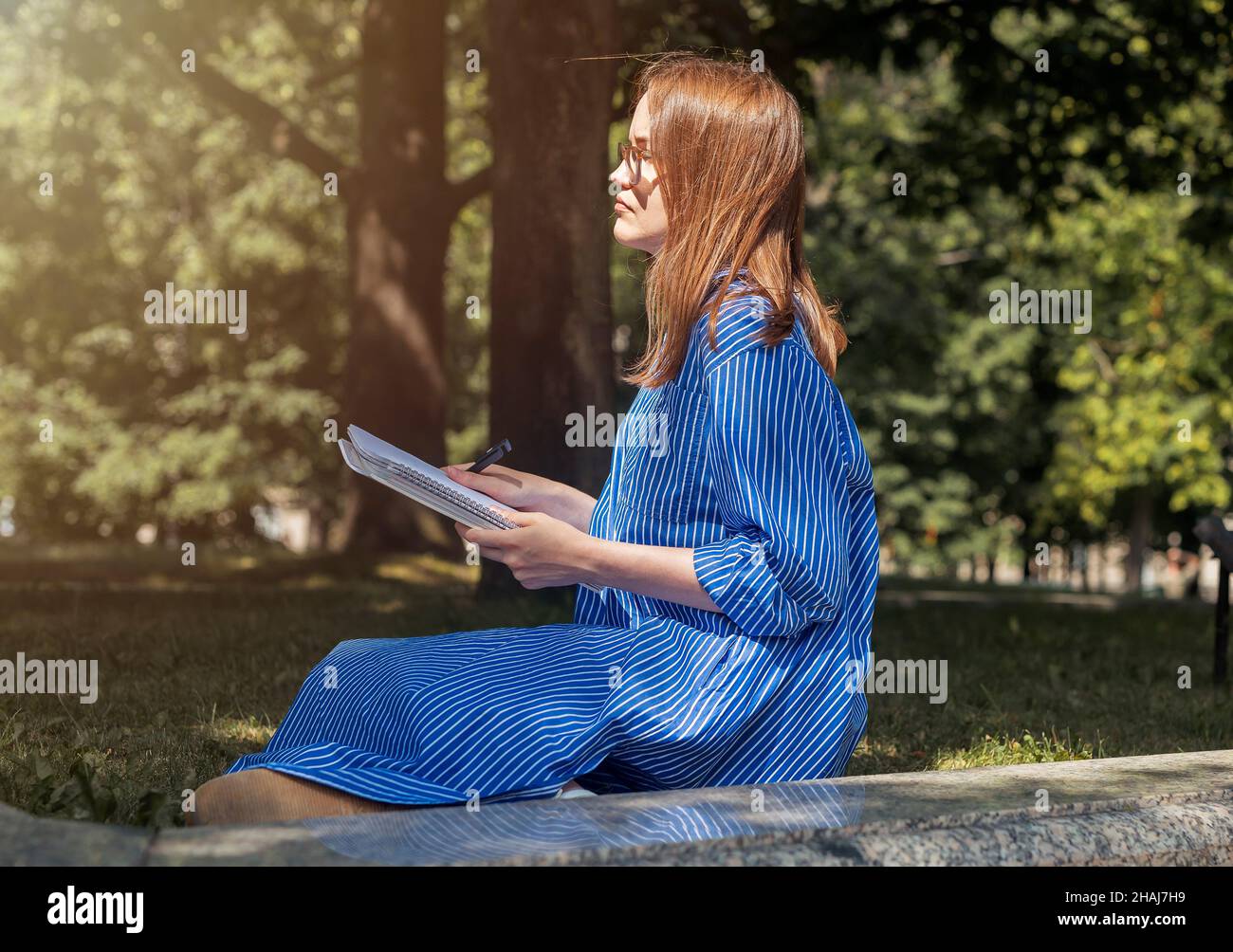Giovane studentessa femminile seduta sull'erba nel parco con taccuino o libro di testo e imparare, leggere e pensare a smth, guardando da parte. Foto Stock