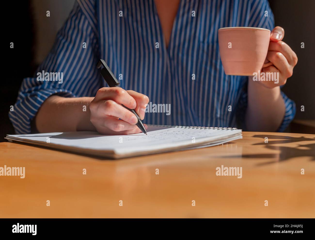 Le mani dello studente scrivono con la penna nel blocco note, prendono appunti e tengono in mano la tazza di caffè. Foto Stock