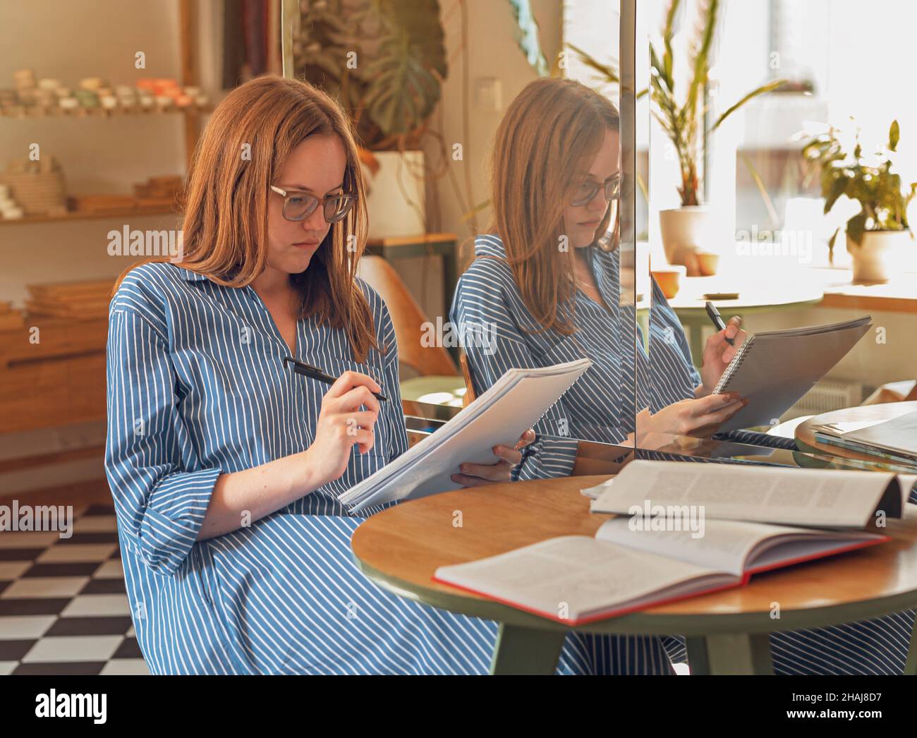 Giovane donna in occhiali in moderno caffè con libri e taccuini, imparare e prepararsi per l'esame. Foto Stock