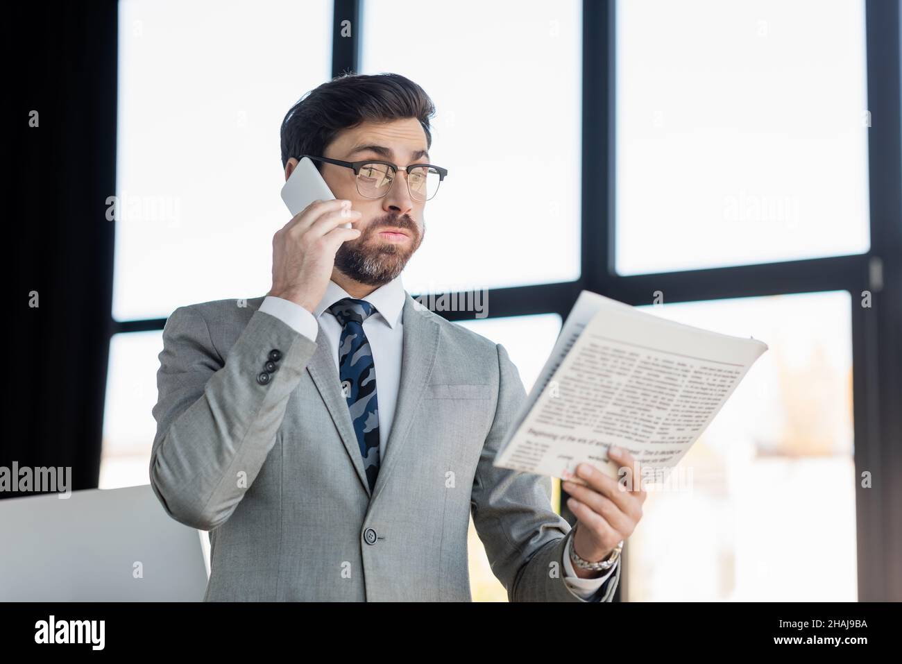 Uomo d'affari che parla sullo smartphone mentre legge le notizie in ufficio Foto Stock