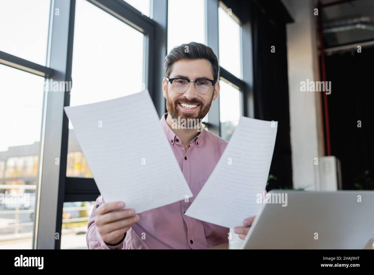 Uomo d'affari sorridente che tiene i documenti vicino al laptop in ufficio Foto Stock