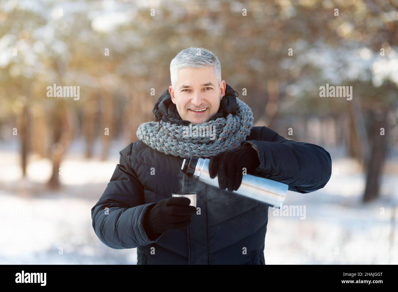 Uomo anziano sorridente in abiti invernali caldi che versano il tè dalla thermos fiasca nel parco innevato Foto Stock