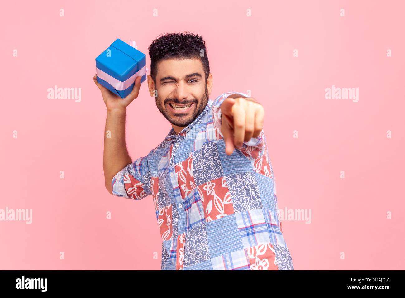 Cattura il tuo regalo. Giovane adulto sorridente uomo vuole buttare fuori la vostra scatola regalo, guardando e puntando la macchina fotografica e winking, indossando la camicia blu. Studio interno girato isolato su sfondo rosa. Foto Stock