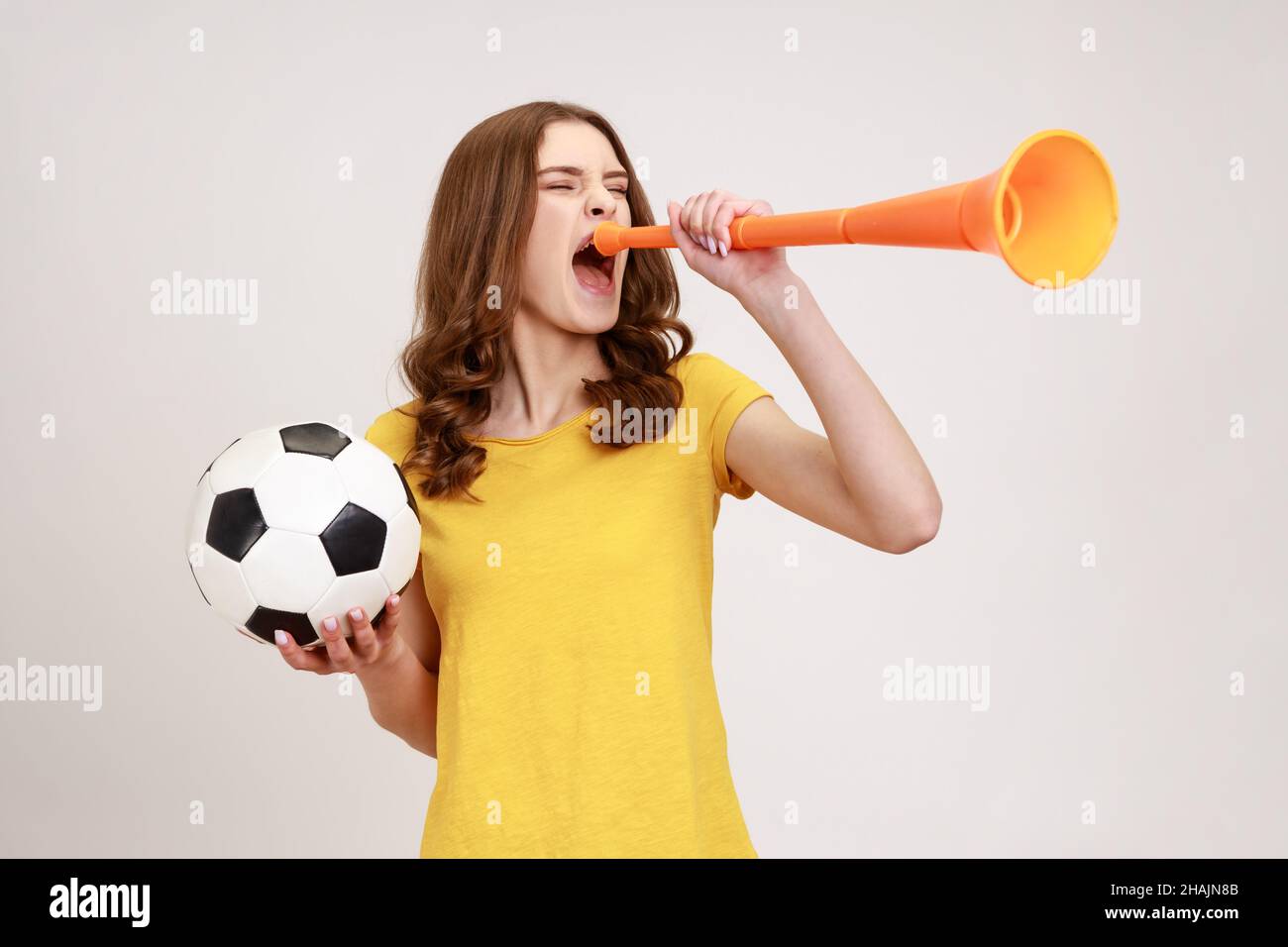 Estremamente positiva ragazza adolescente soddisfatto in giallo t-shirt urlando in corno che tiene palla di calcio, celebrando la vittoria della squadra di calcio preferita. Studio interno girato isolato su sfondo grigio. Foto Stock