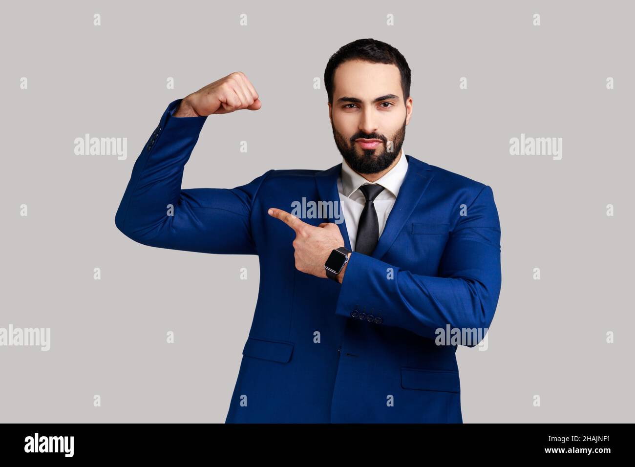 L'uomo robusto bearded alza le mani mostrando il potere, indicando il suo bicipite, sentendosi indipendente forte con il look orgoglioso, indossando il vestito di stile ufficiale. Studio interno girato isolato su sfondo grigio. Foto Stock