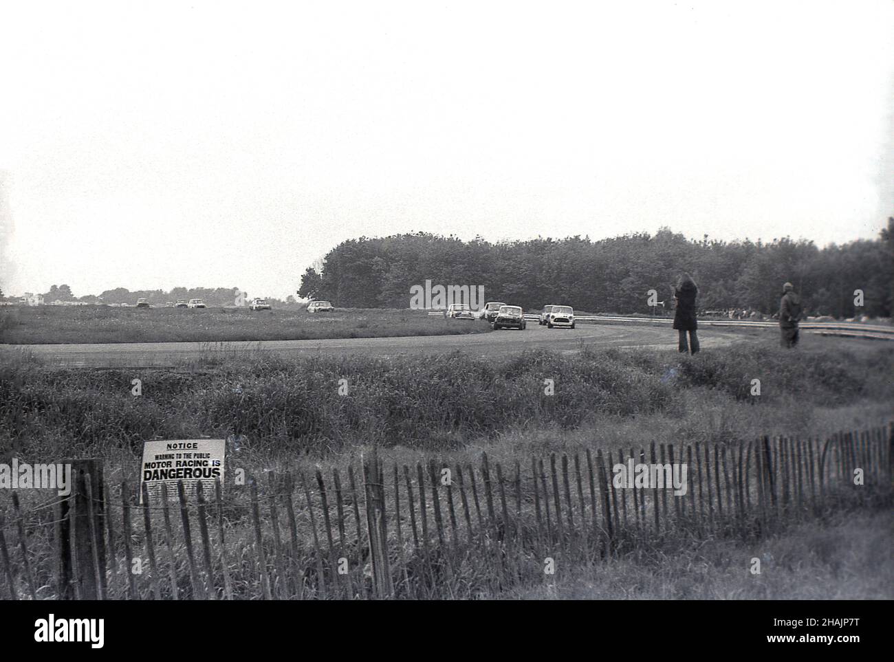 1960s, storico, motorsport, auto da corsa, Inghilterra, REGNO UNITO. Una donna e un uomo si levano in piedi sull'erba accanto alla pista. Un piccolo avviso sull'erba da una breve recinzione in legno dice, Motor Racing è PERICOLOSO. Foto Stock