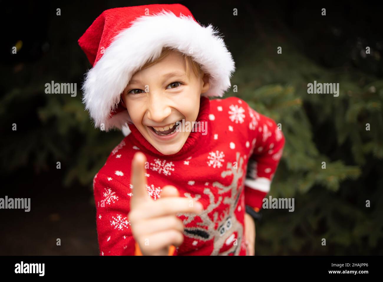 Un ragazzo in un maglione di Natale a maglia rossa con una renna di Natale e un cappello di Babbo Natale onderà il dito indice alla macchina fotografica e ride Foto Stock