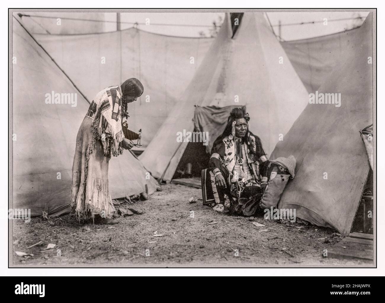 Papoose North American indigena indiana americana donna della regione del Northwest Plateau, scattando una fotografia con una pellicola Kodak mantice mano fotocamera di un uomo e un bambino in una culleboard di fronte a tipis. La gente è probabilmente esecutori in uno spettacolo del selvaggio West o in uno spettacolo laterale. 1911 - Indiani d'America del Nord--Abbigliamento & vestire--Northwest, Pacifico--1910-1920 telecamere--1910-1920 Tipis--1910-1920 stampe in gelatina d'argento--1910-1920. Foto ritratto--1910-1920. Foto Stock