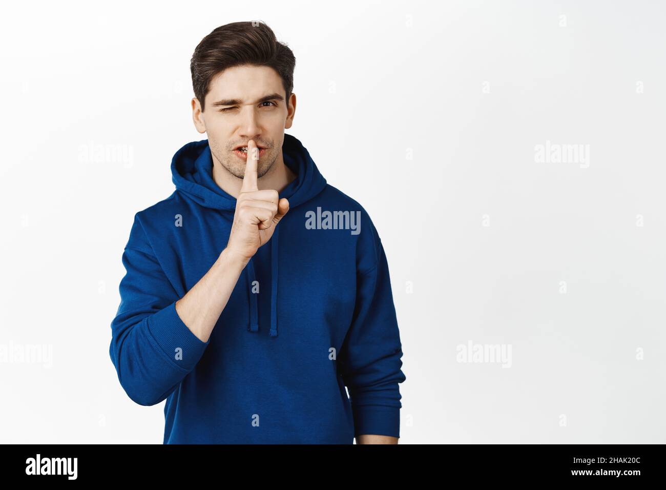 Bel ragazzo flirty winking, mostrando hush, shush non parlare gesto e winking, ha segreto, essere silenzioso segno, in piedi su sfondo bianco Foto Stock