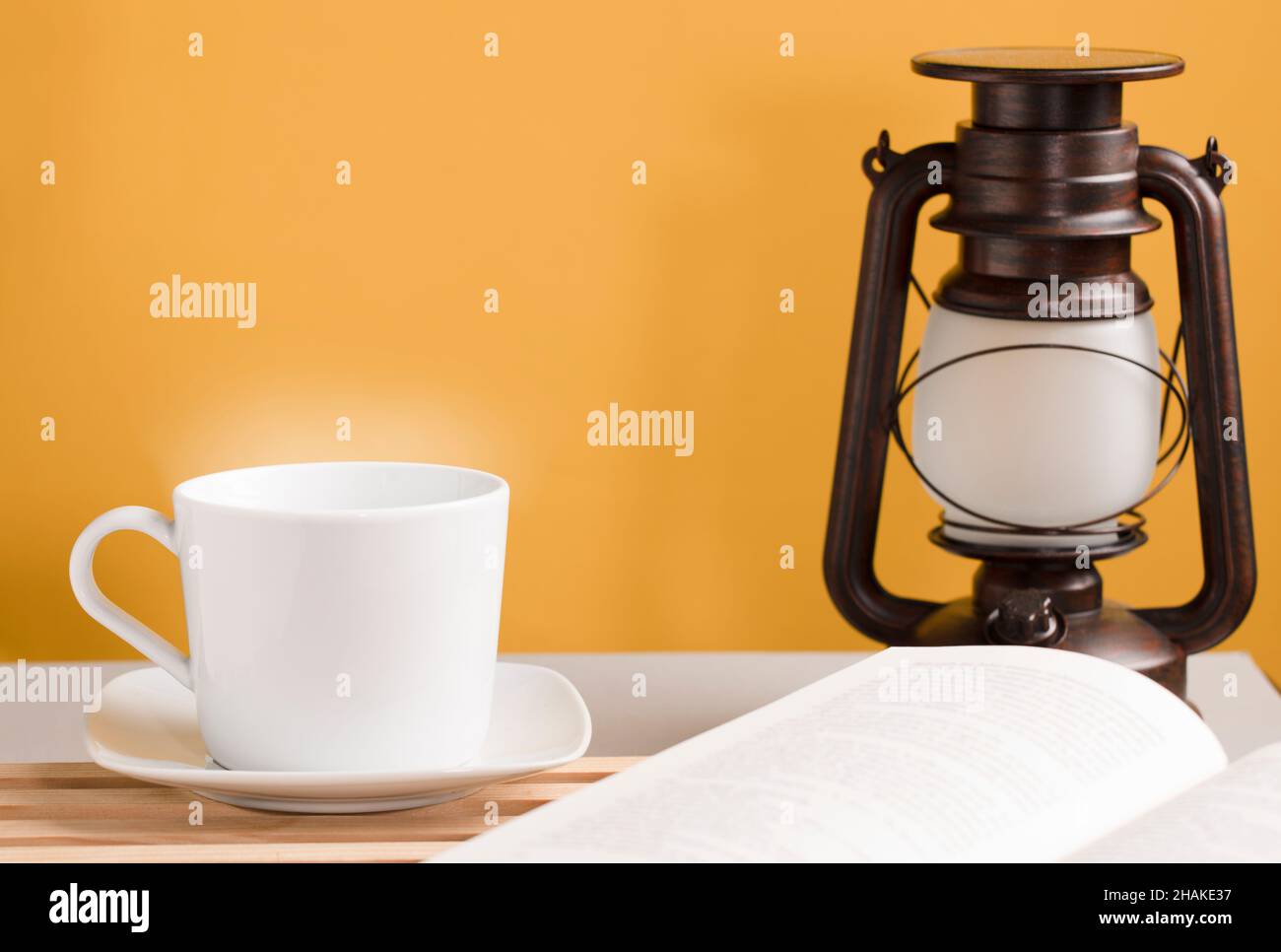 Tazza di caffè bianca con vapore caldo, sulla scrivania con libro aperto e lampada solare, sfondo giallo-arancione Foto Stock