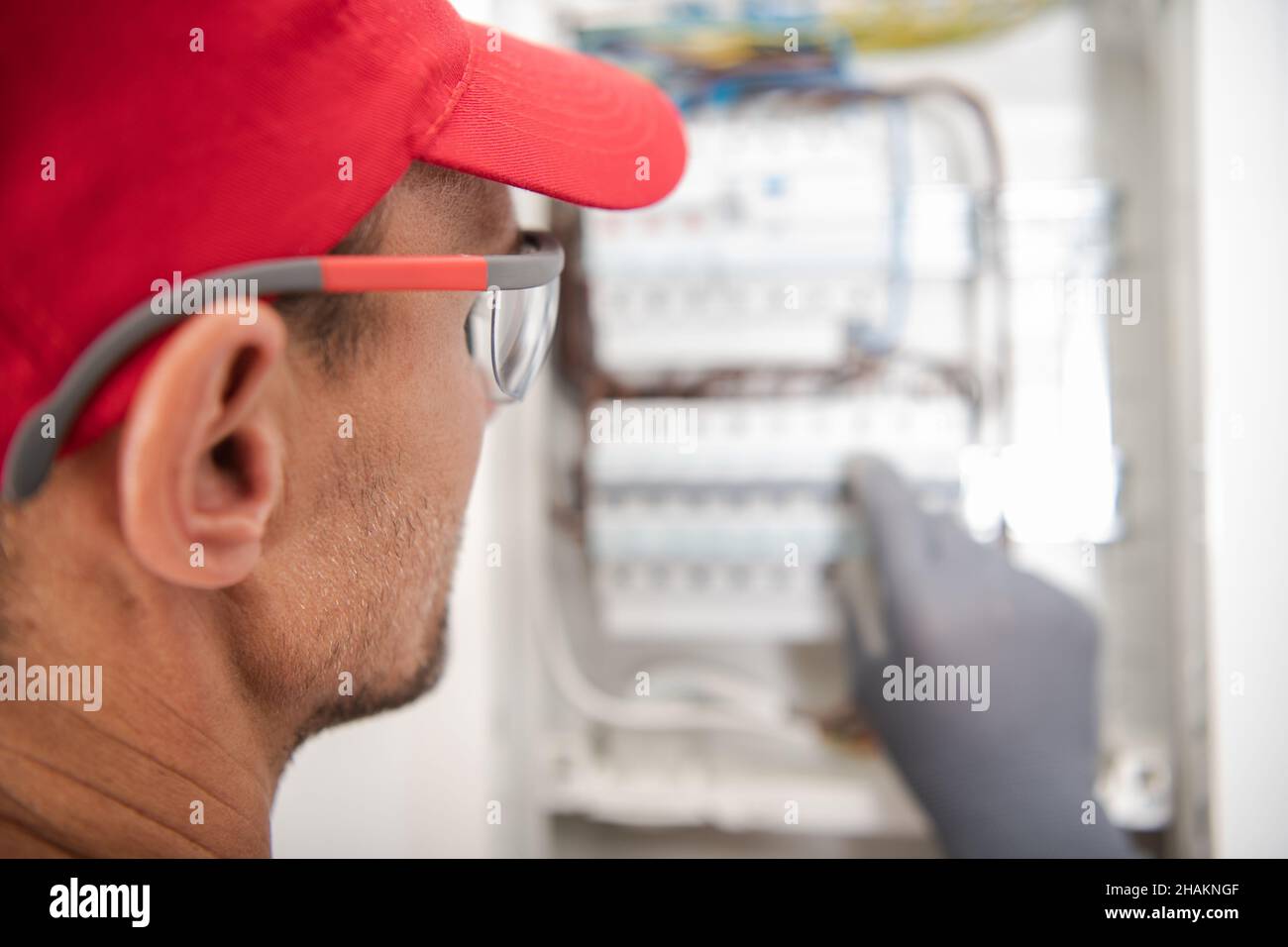 Gli elettricisti caucasici indossano occhiali di sicurezza che guardano all'interno della scheda di interruzione elettrica per potenziali problemi. Circuito elettrico residenziale. Foto Stock
