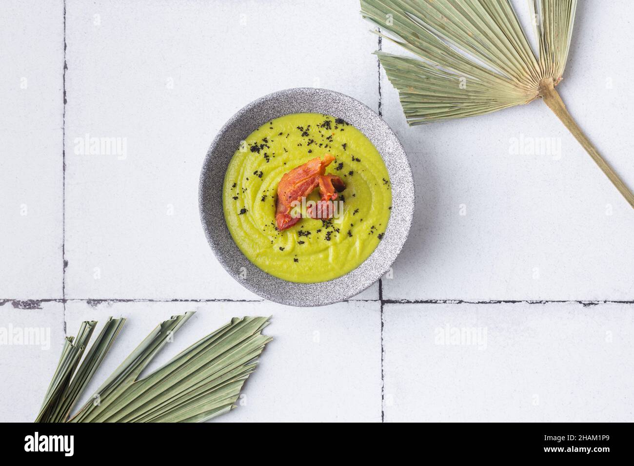 Purea di zuppa verde con pancetta alla griglia su sfondo piastrellato foglie di palma Foto Stock