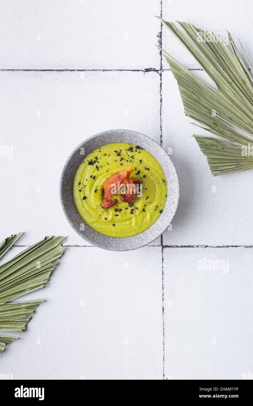 Purea di zuppa verde con pancetta alla griglia su sfondo piastrellato foglie di palma Foto Stock