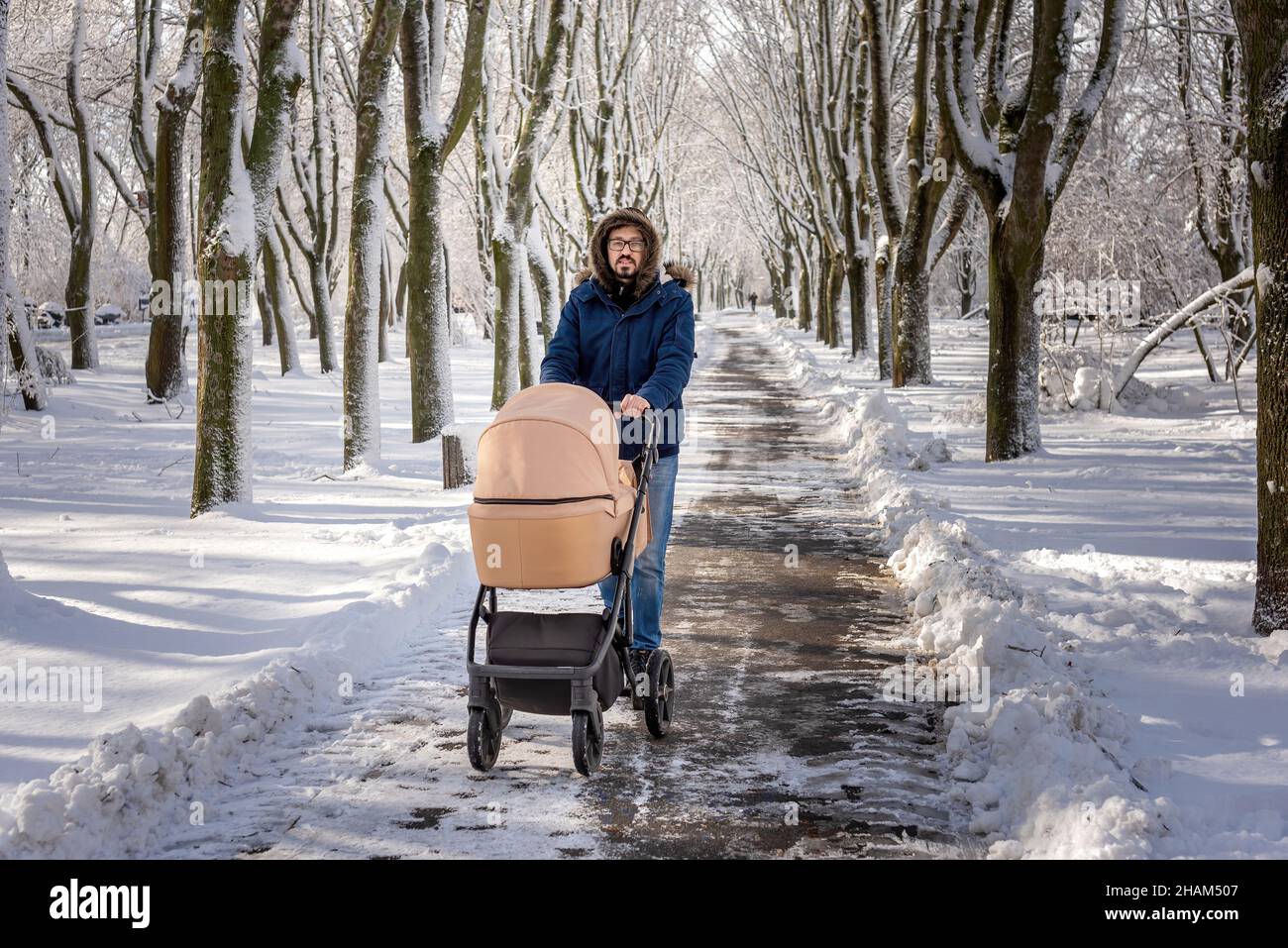 Padre che cammina con passeggino beige in un parco coperto di neve. Freddo bellissimo giorno d'inverno. Passeggiate ricreative con un bambino. Uomo in giacca e jeans in anorak blu. Foto Stock