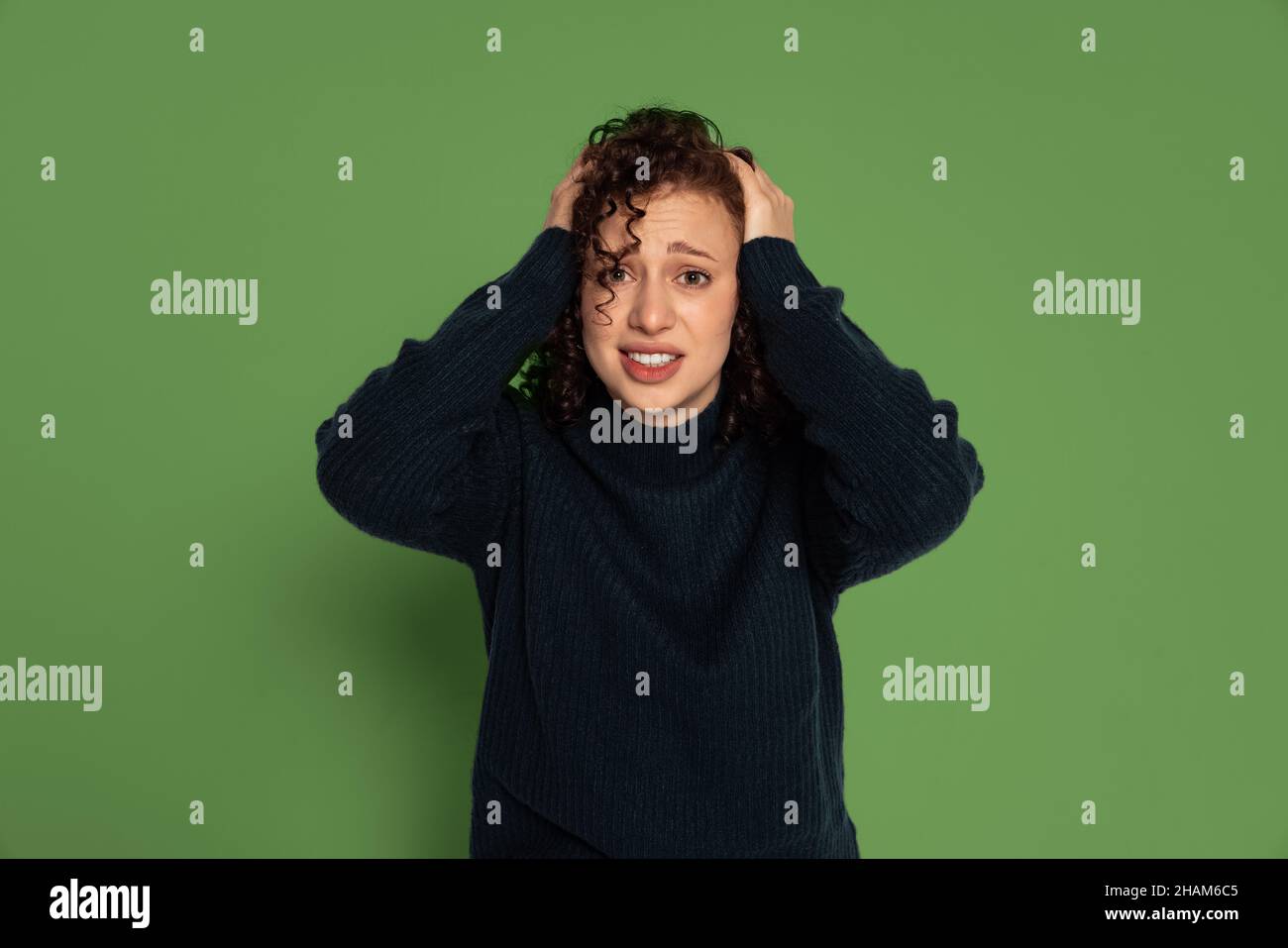 Una giovane ragazza emotiva in caldo maglione invernale che tiene la testa con le braccia isolate su sfondo verde studio. Concetto di emozioni Foto Stock