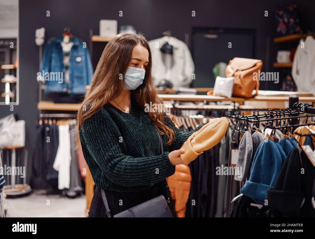 Giovane donna che indossa maschera facciale facendo acquisti stagionali di vestiti per i membri della famiglia, guardando caldo cappello a maglia nelle sue mani mentre si trova in piedi nel reparto moda del centro commerciale Foto Stock